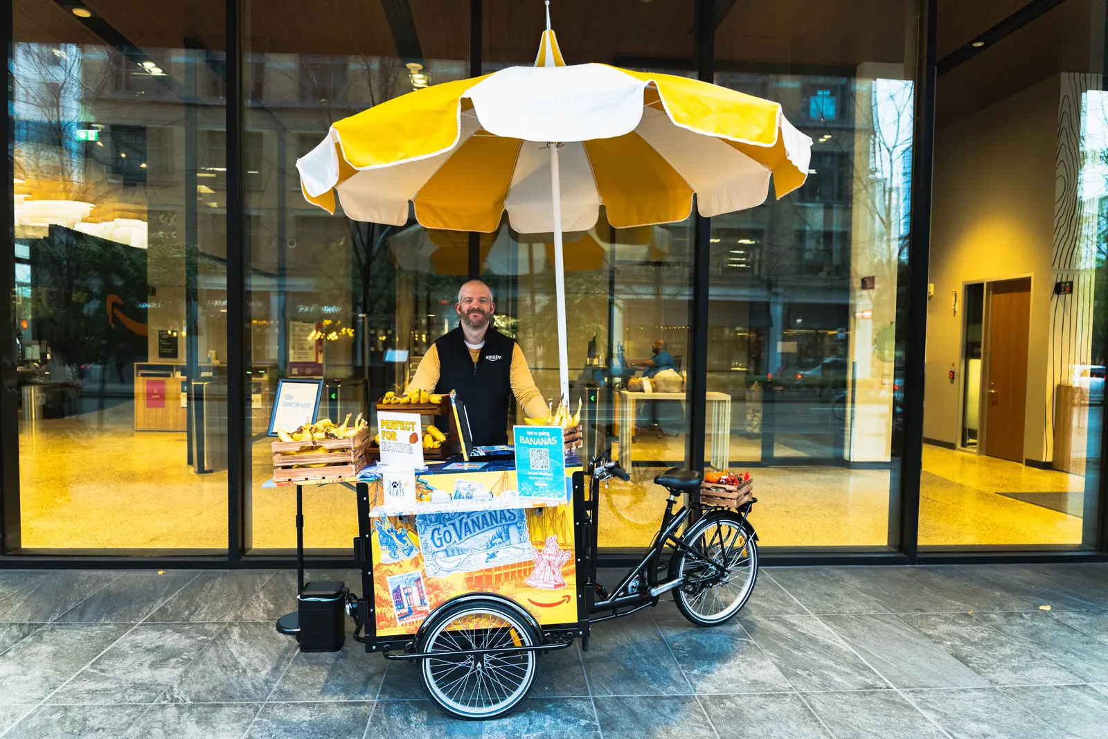 A person in a dark vest stands behind a bicycle cart stocked with bananas under a yellow-and-white striped umbrella, in front of a glass office building entrance