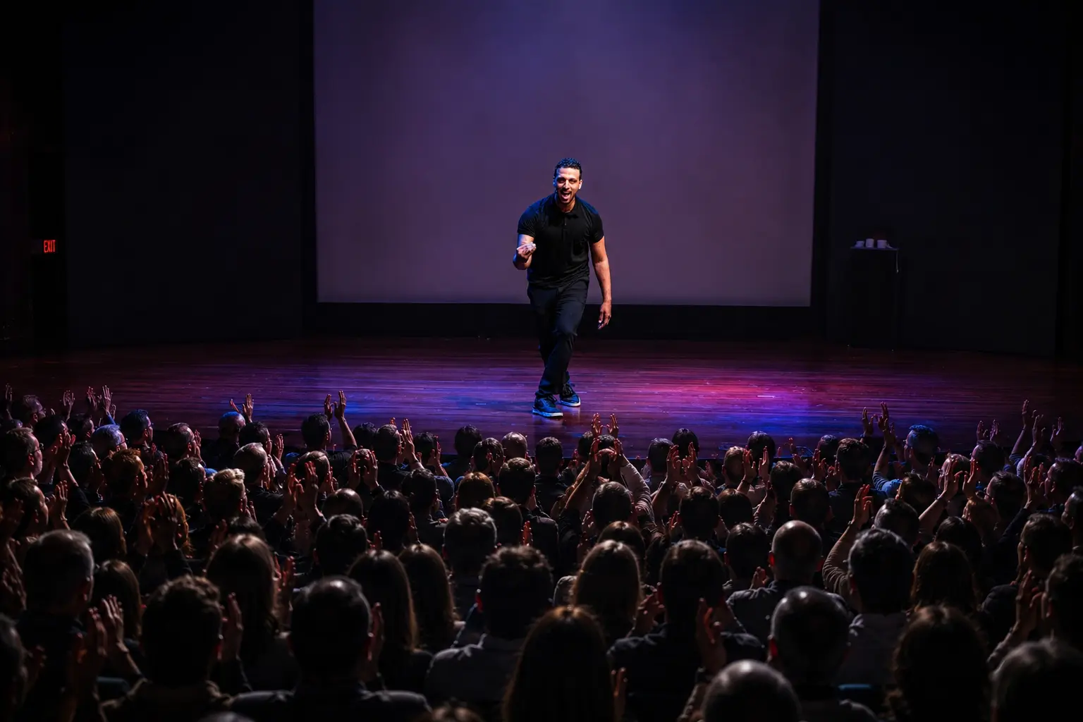 A performer stands on a lit stage addressing a seated audience, with a large screen displayed behind him.