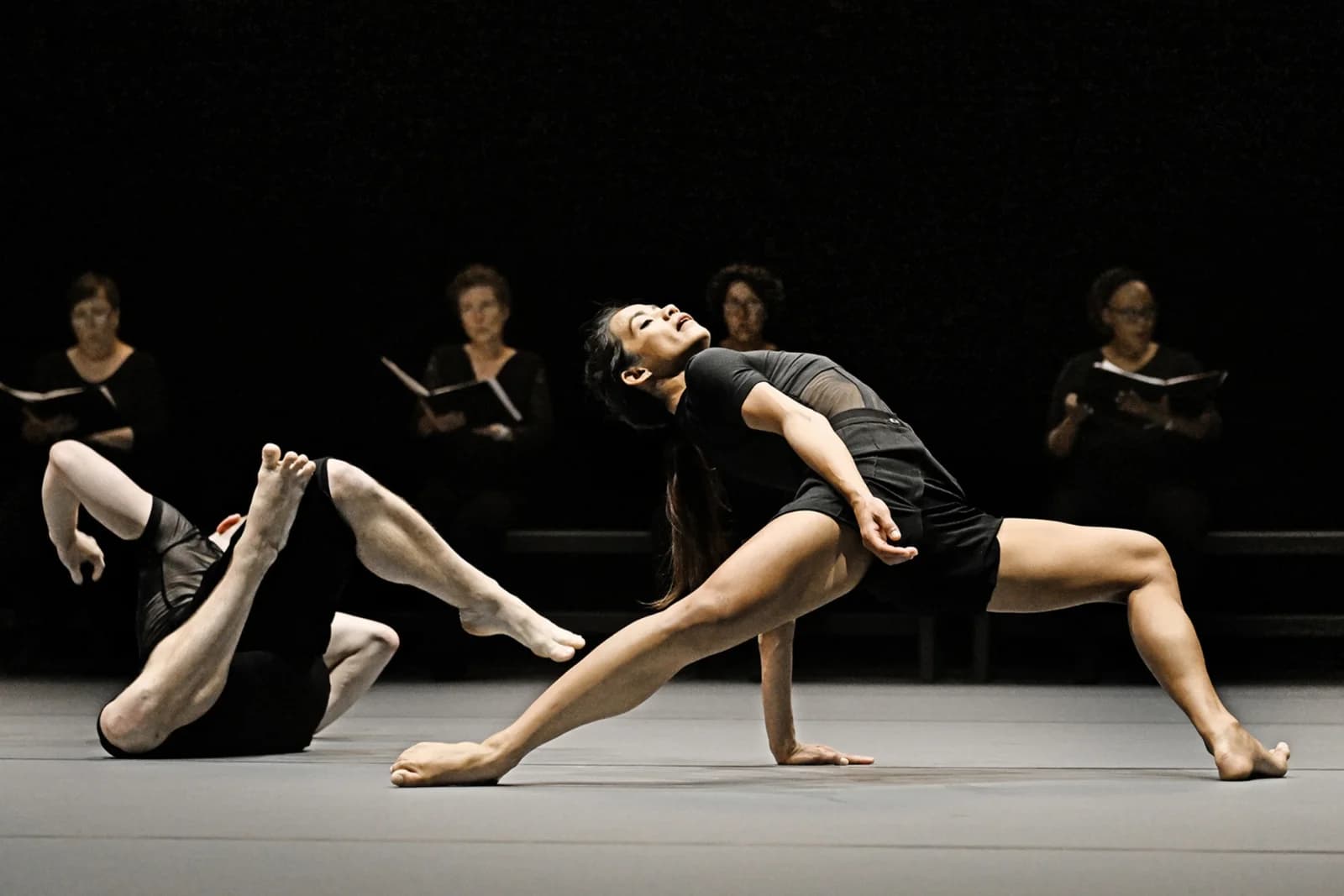 Two dancers on a bare stage with choir singers seated behind them, in an earlier staging of I'm afraid to forget your smile, as published by Ballet BC