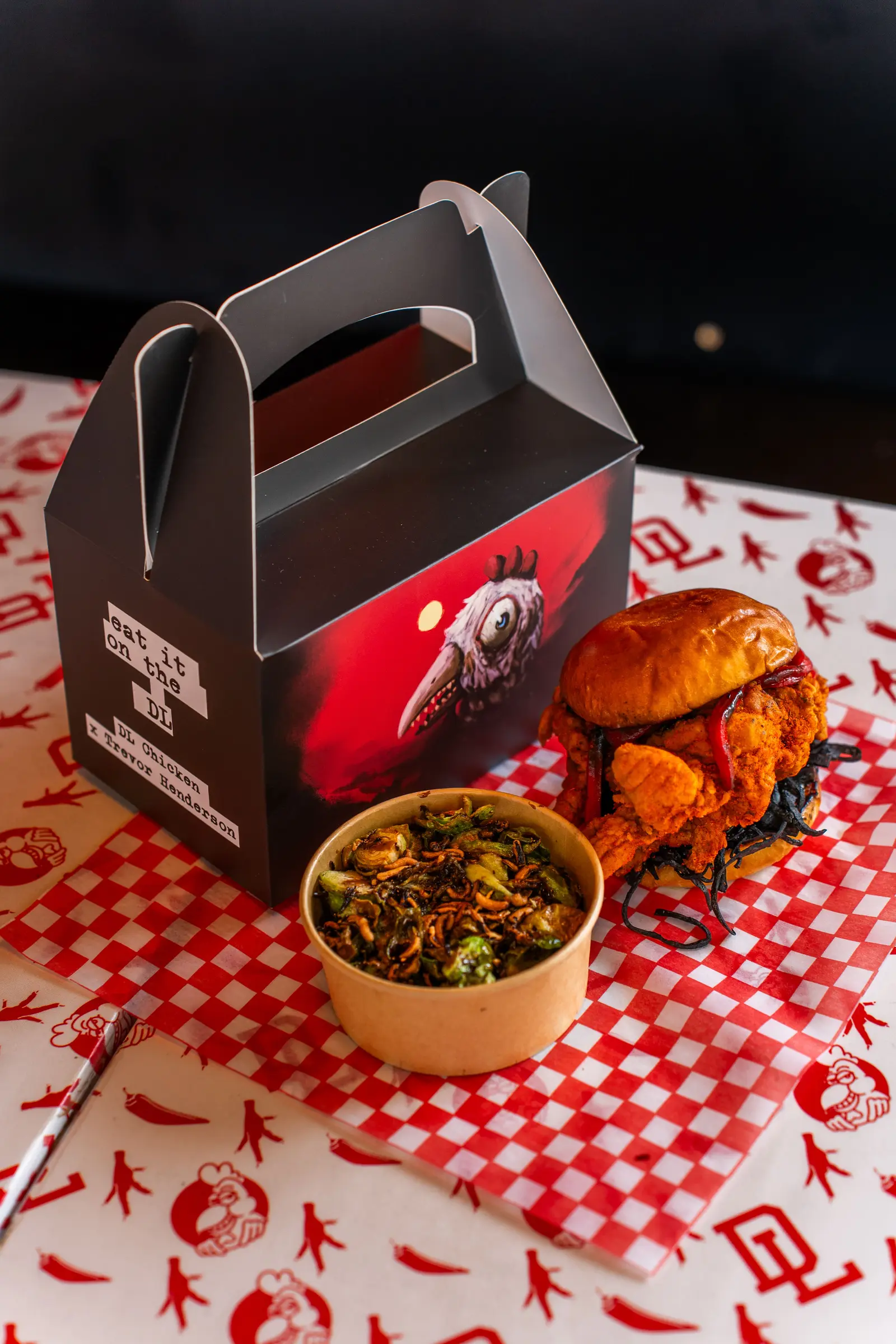 A fried chicken sandwich, a bowl of greens, and a black-handled takeout box from DL Chicken's Halloween collaboration menu, arranged on red-and-white checkered paper.