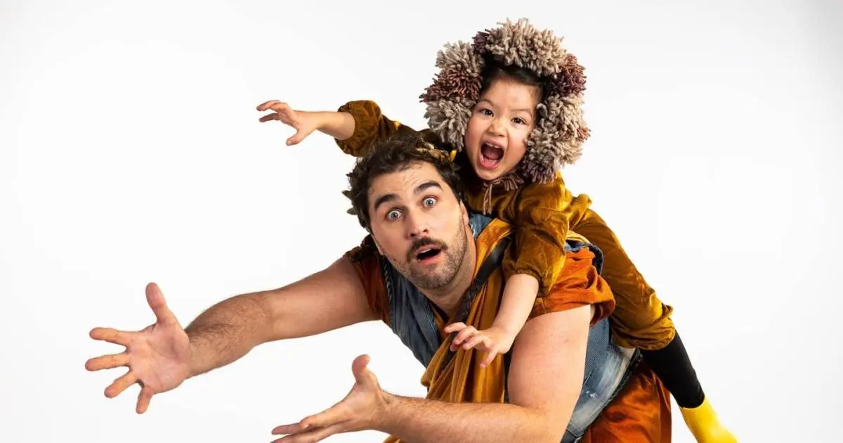 A performer in a colourful patterned costume leans forward, arms outstretched, a curly-haired child on their shoulders, both mouths wide open