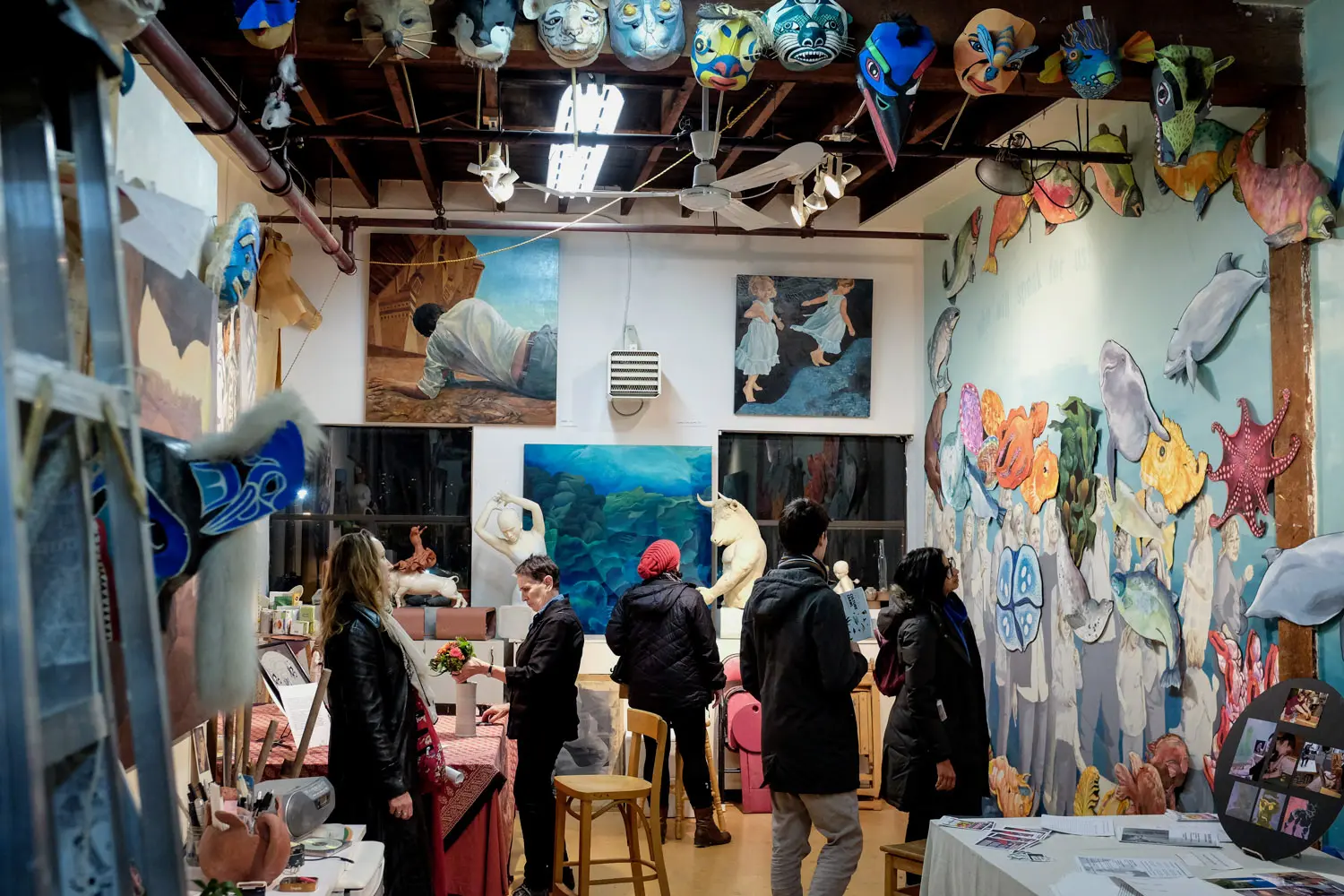 Visitors inside an artist's studio with painted fish sculptures hanging overhead, paintings on a white wall and a mural on the right