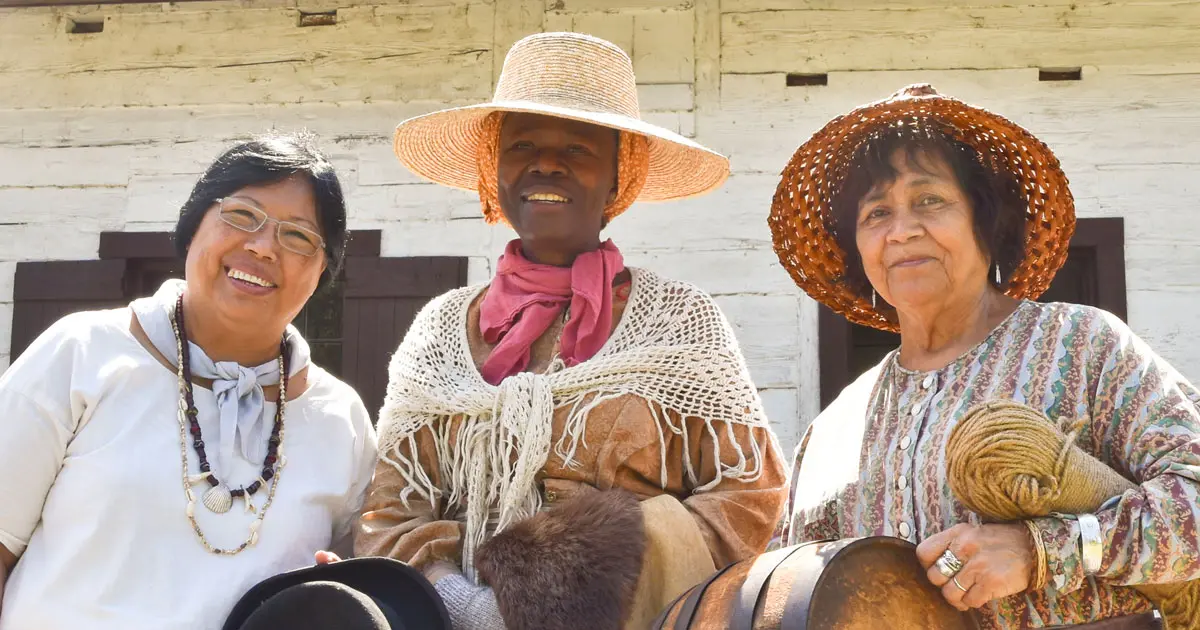 Three people in period-style clothing and hats stand in front of a weathered white wooden wall at Fort Langley National Historic Site.