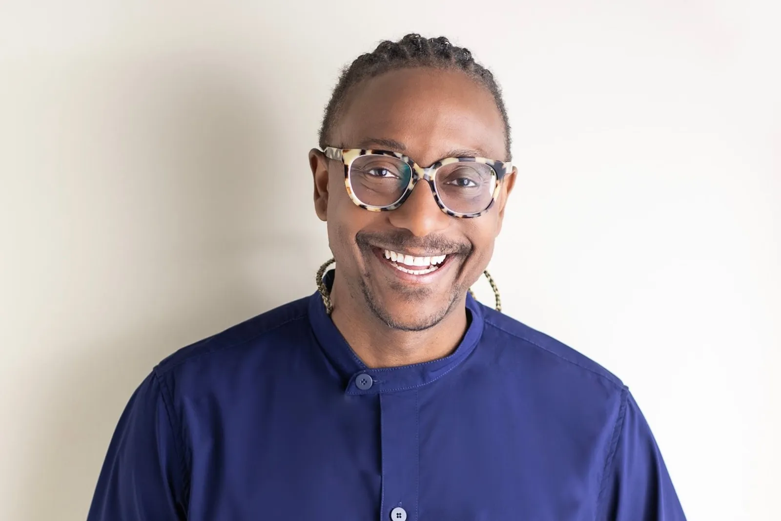 Portrait of chef Gregory Gourdet smiling, wearing patterned glasses and a navy shirt, against a pale wall