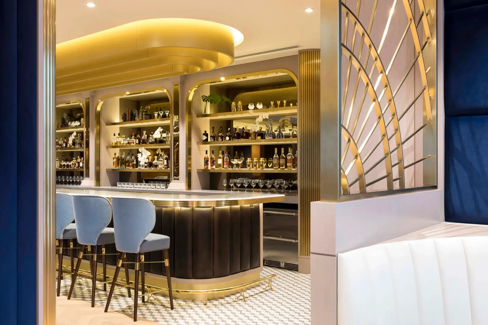 A curved dark bar with gold trim in front of illuminated gold-toned shelves of bottles and glassware, with pale blue upholstered bar stools