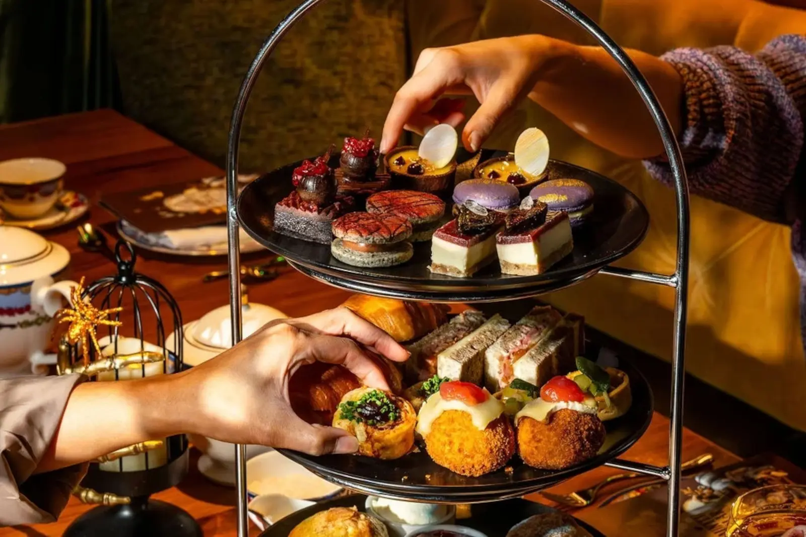 Three-tiered afternoon tea service with handcrafted pastries, savoury bites and gothic-themed desserts; two hands select items from the display