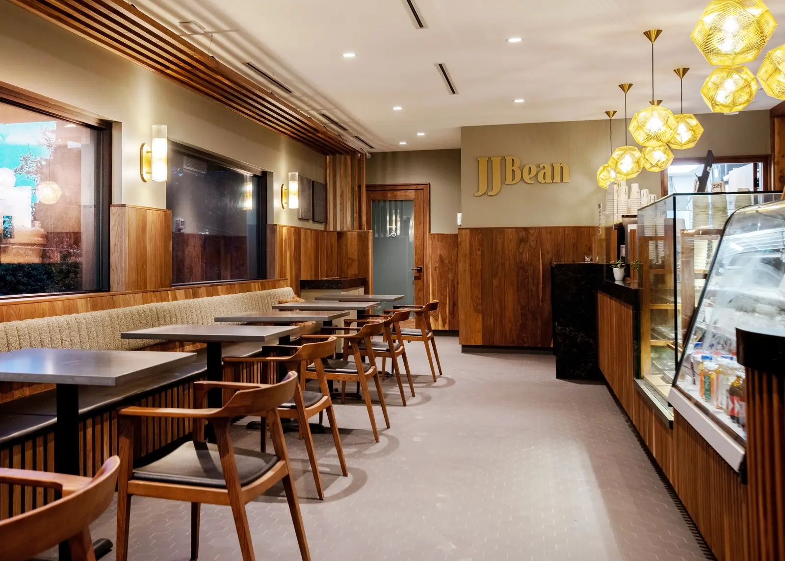 Interior of an existing JJ Bean cafe, not the new Broadway location, with wood-panelled walls, booth seating, wooden chairs, globe pendant lights and a pastry case