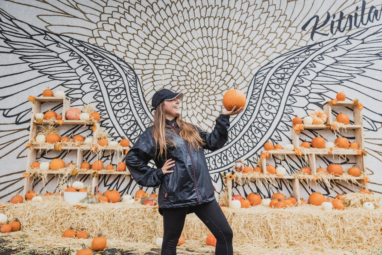 A person in a dark jacket and cap holds up a pumpkin in front of a large black-and-white wing mural, with pumpkins on wooden crates and hay bales nearby