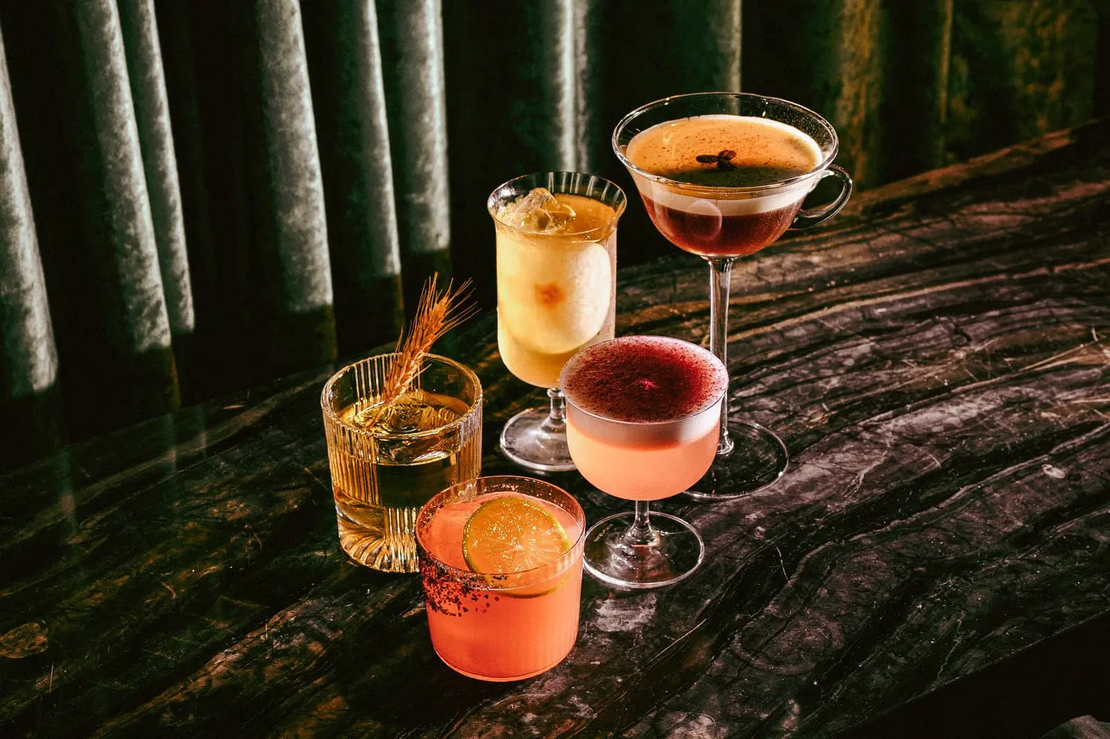Five colorful cocktails arranged on a dark, textured tabletop against a dim, draped background at Prophecy bar.