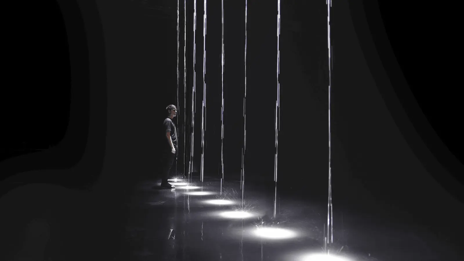 A person stands in a dark room beside tall, thin vertical strands of light, with bright pools of white light on the floor.