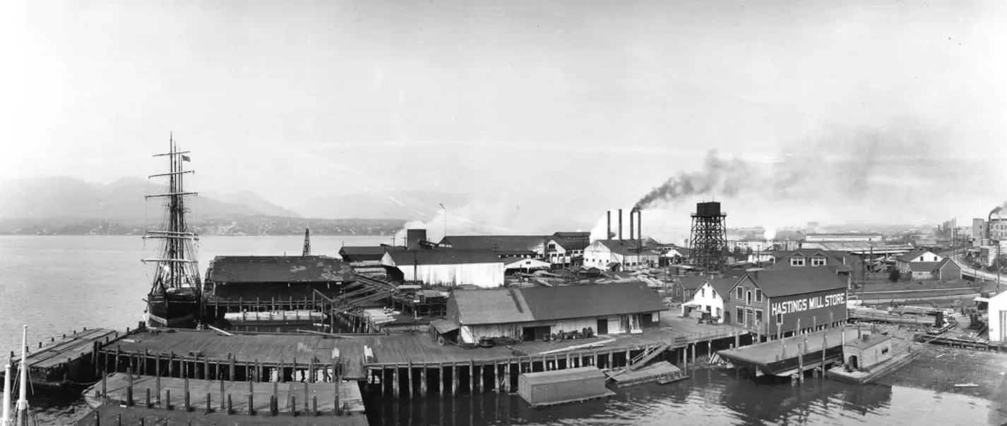Archival black-and-white panorama of a historic waterfront with a tall-masted sailing ship, wooden wharves, smoking stacks and a building signed Hastings Mill Store
