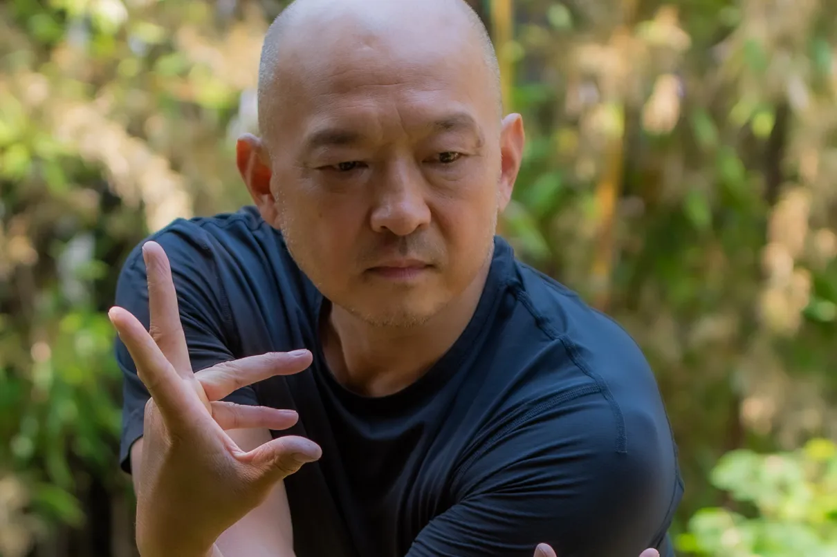 Choreographer Wen Wei Wang, in a dark T-shirt, holds an expressive pose with his hands and arms crossed in front of a leafy garden