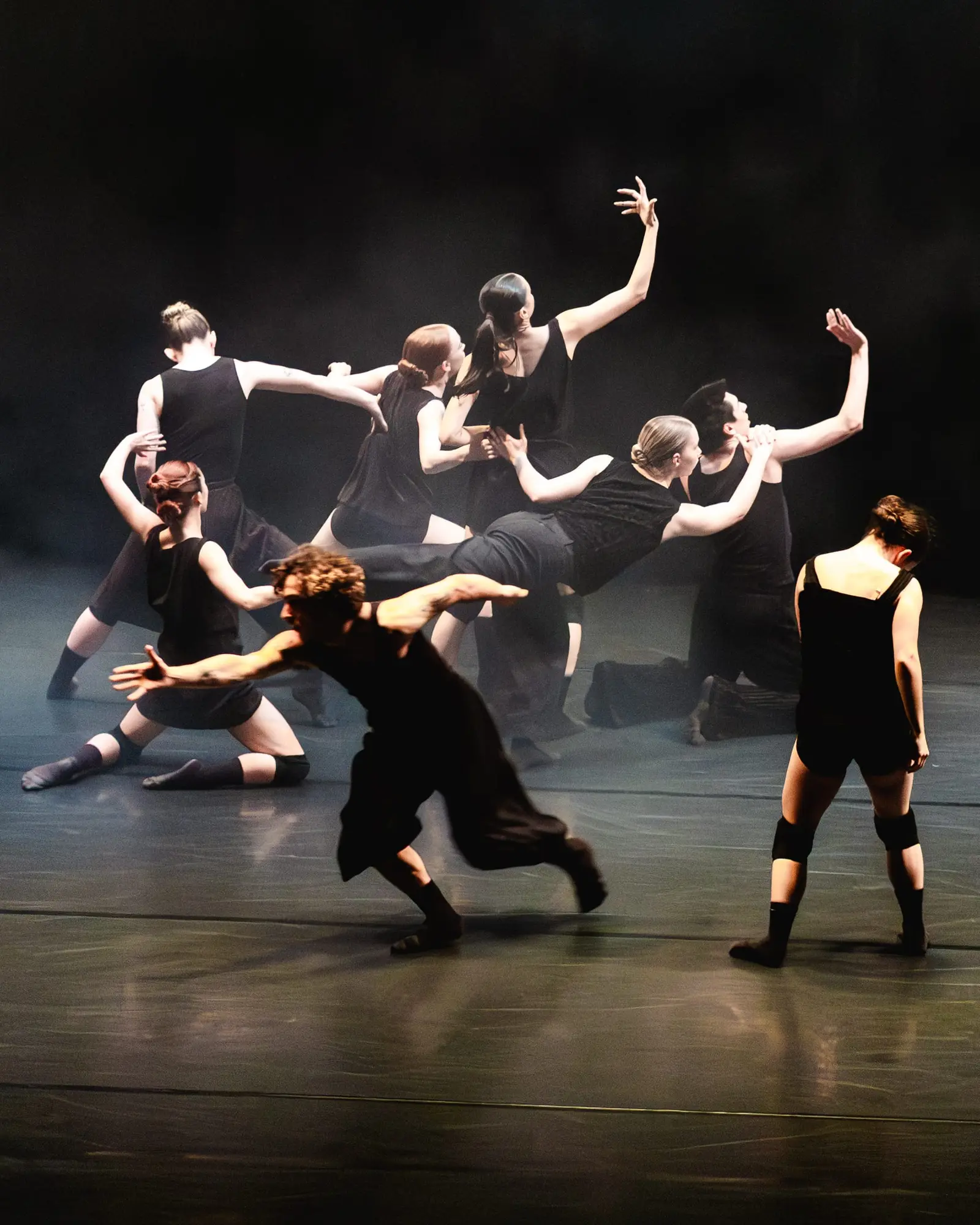 Several dancers in dark and light clothing perform together on a dimly lit stage, with some seated or crouched and others extending their arms.