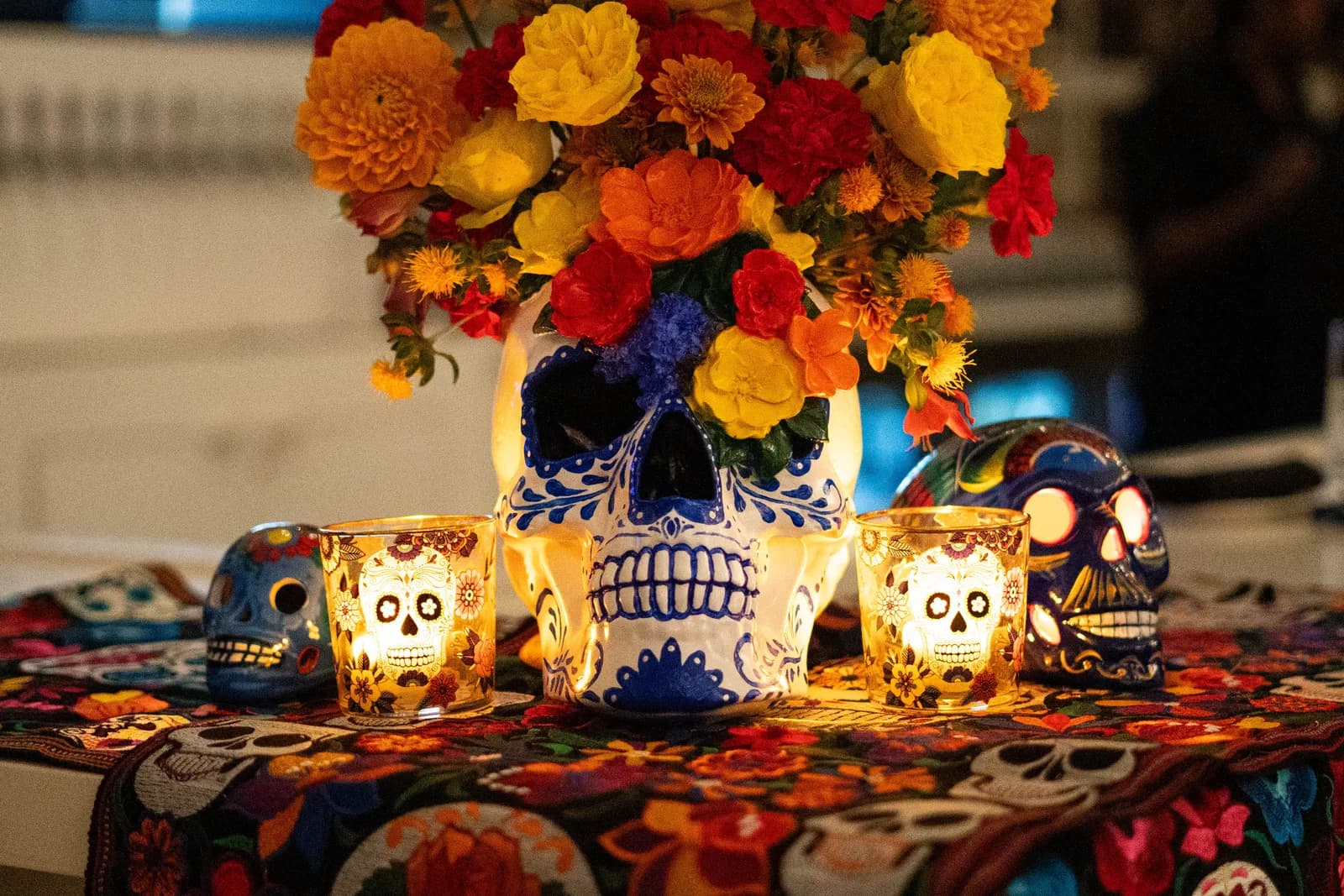 Día de los Muertos table decor on Botanist's event page: a painted sugar skull vase of marigolds and roses beside skull candle holders on an embroidered cloth