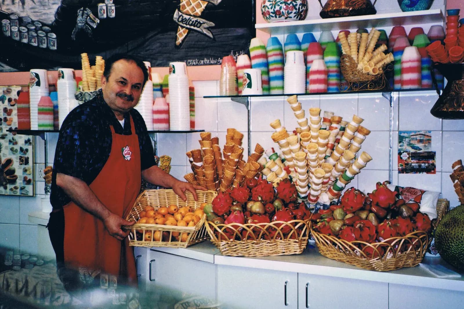 Behind the counter at La Casa Gelato, with stacked waffle cones, striped cups and baskets of fresh fruit on display