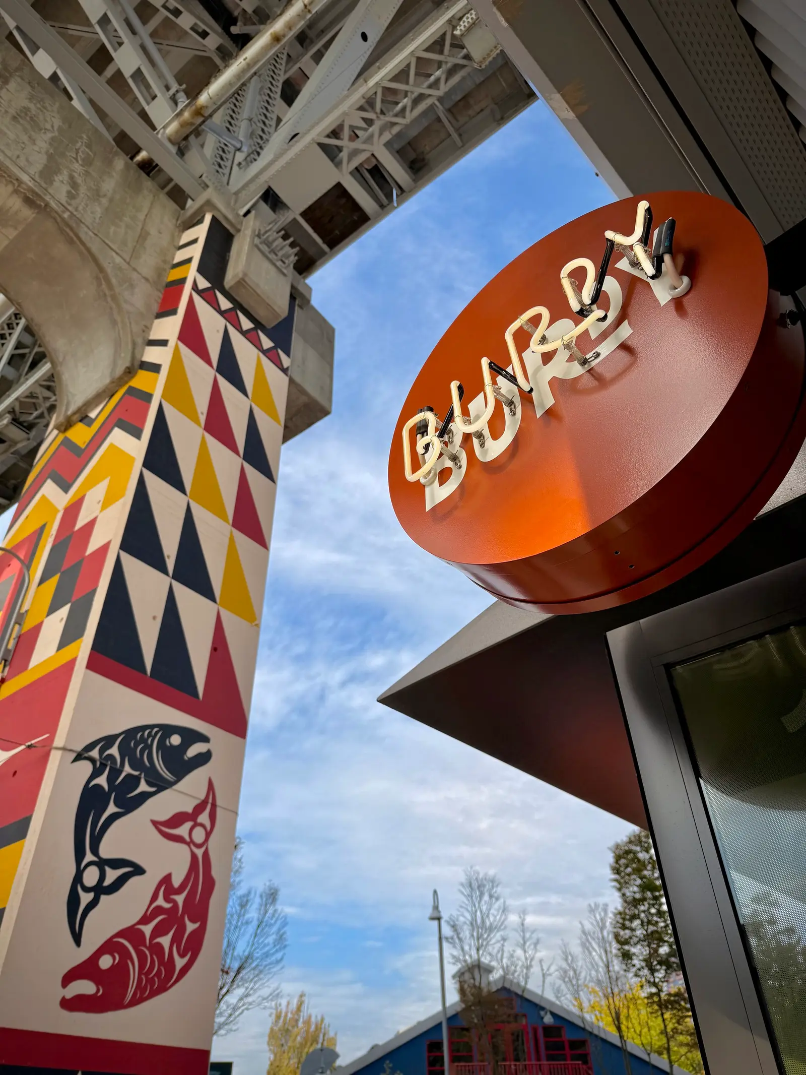 A round orange sign with white lettering hangs above the Burdy restaurant entrance beside a colorful geometric mural and glass windows on Granville Island.