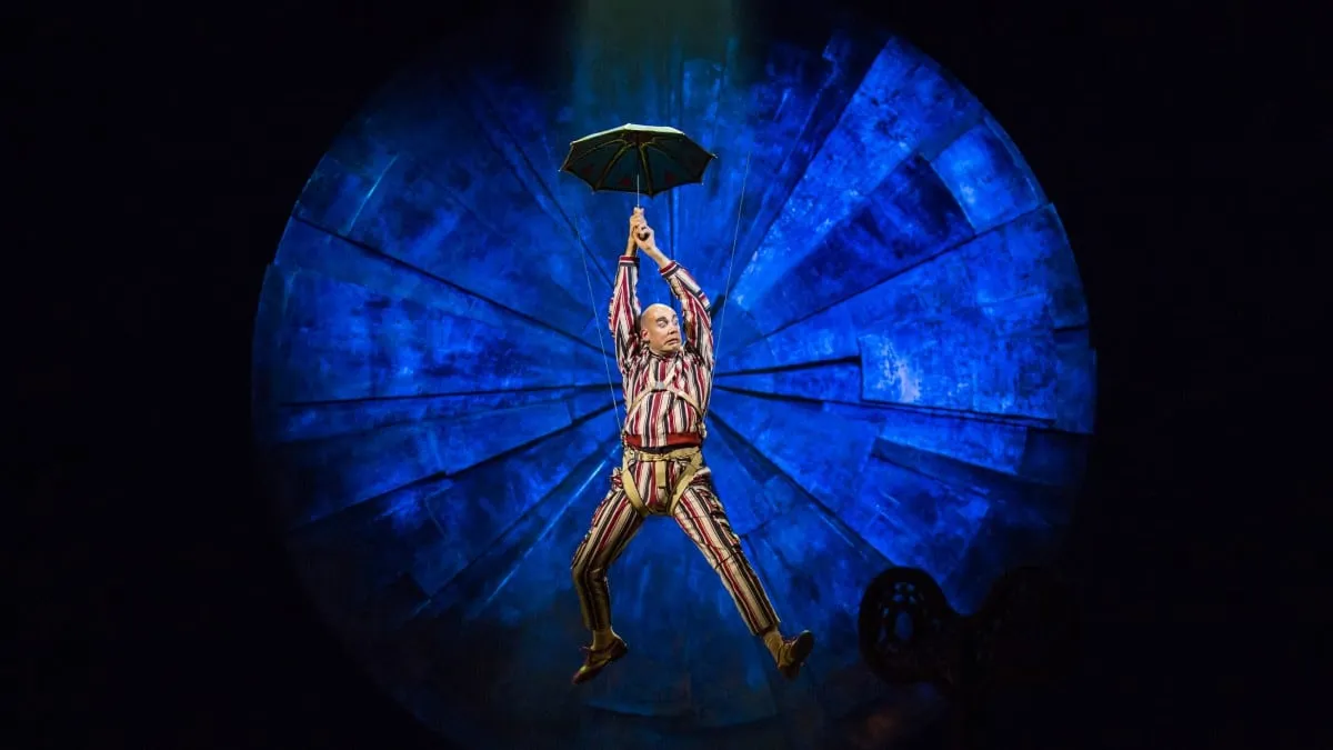A LUZIA performer in a striped suit floats down holding a small umbrella in front of a glowing blue circular backdrop
