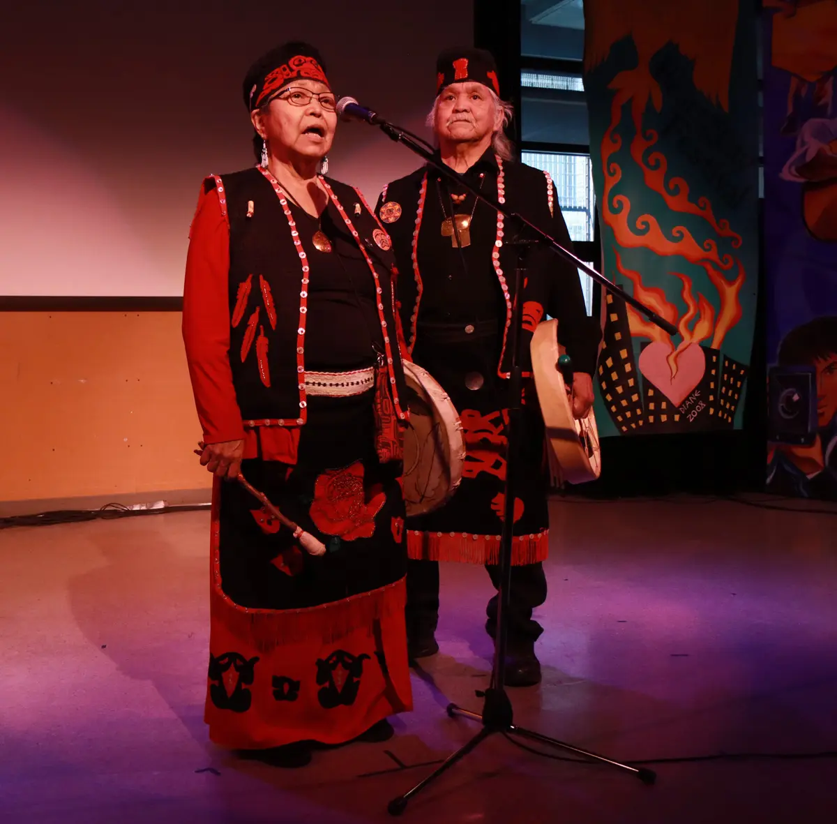 Two performers in black and red clothing stand at microphones on a lit stage, from a previous year's Heart of the City Festival opening ceremony