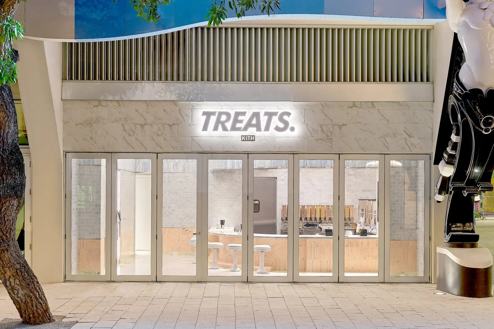 Exterior of a Kith Treats shop with the word TREATS on a light marble facade, glass double doors, a blue and white awning and a tree at left