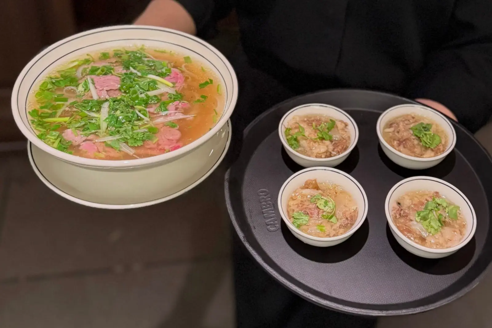 A large bowl of pho with herbs and sliced meat beside a tray holding four smaller bowls of noodle soup on a dark tabletop.