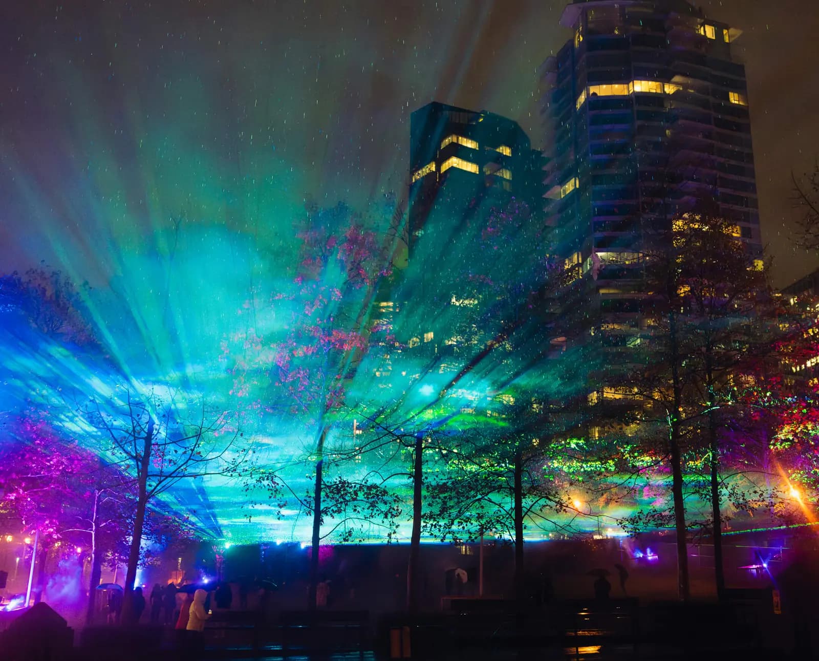 Blue, green, purple and pink light beams cross the night sky above bare trees, with lit office towers behind and silhouetted people below, at a previous Lumiere Festival