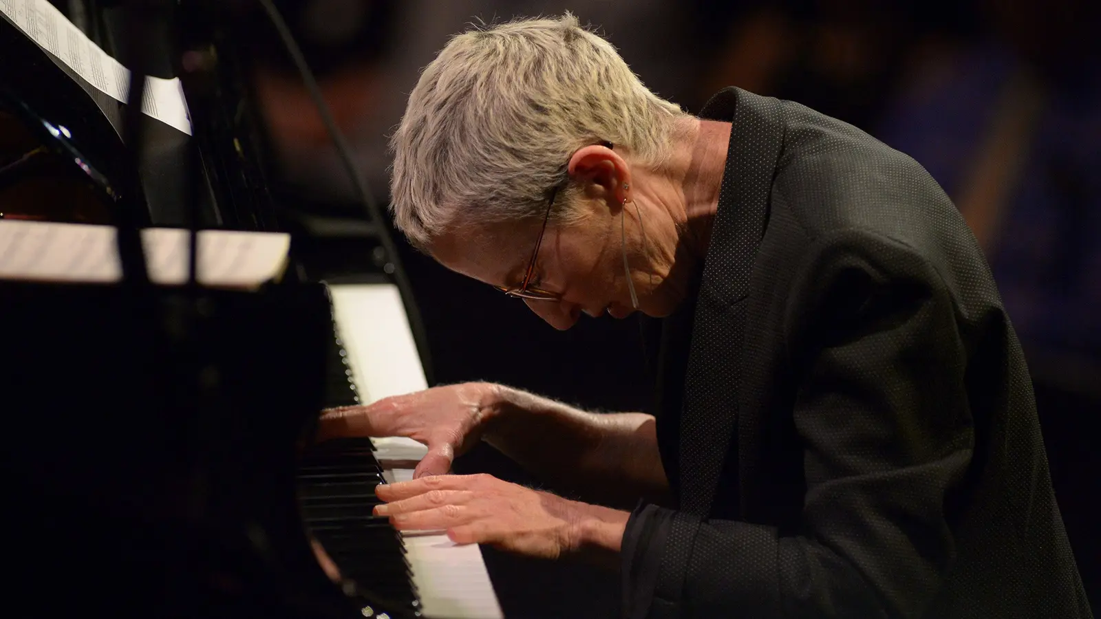 A pianist with short light-coloured hair, glasses and a dark jacket leans over a piano keyboard on a dark stage
