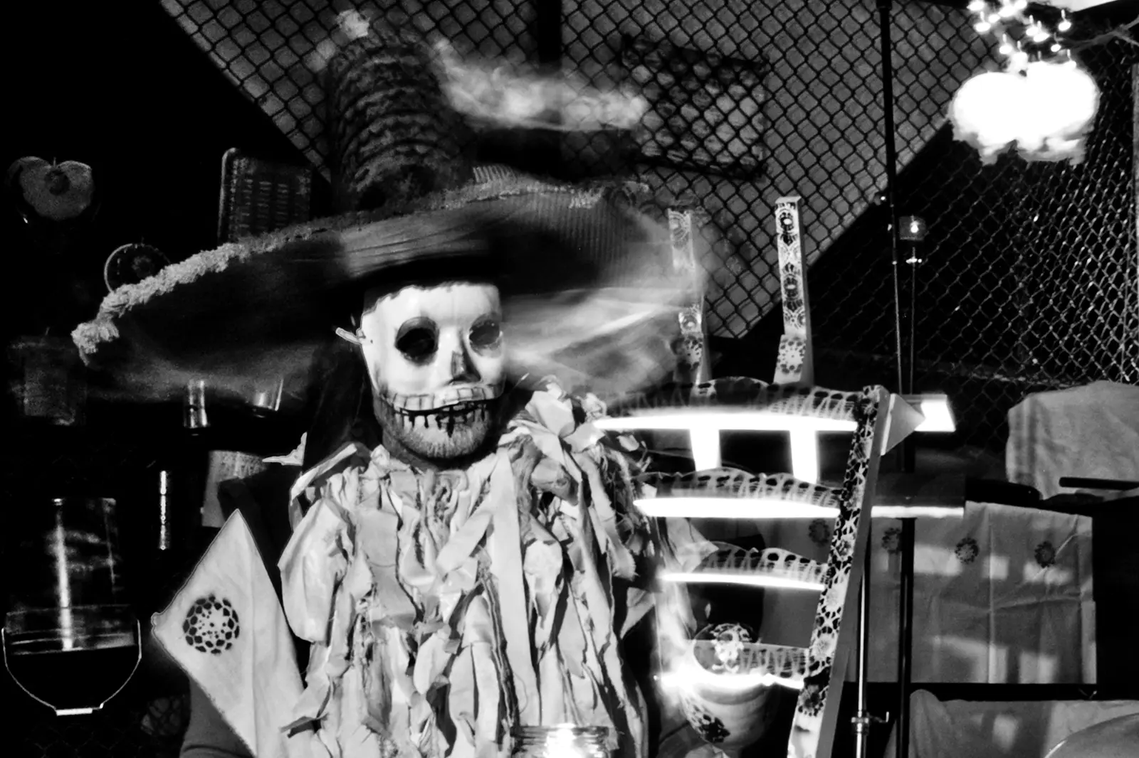 Black and white photo of a performer in a skull mask and wide sombrero beside candles and jars, from the organiser's Parade of Lost Souls page