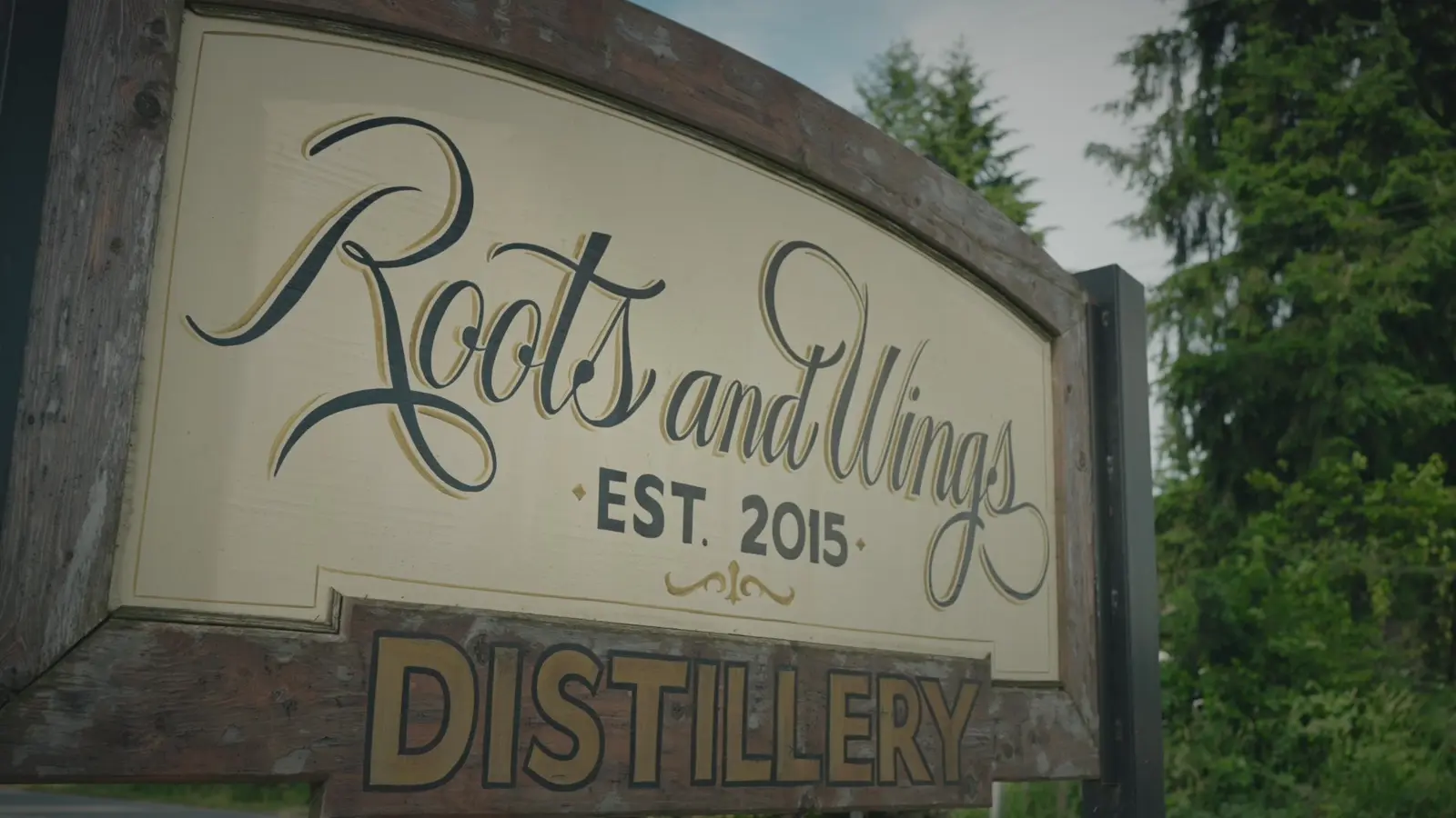 A large Roots and Wings Distillery sign mounted outdoors among trees.