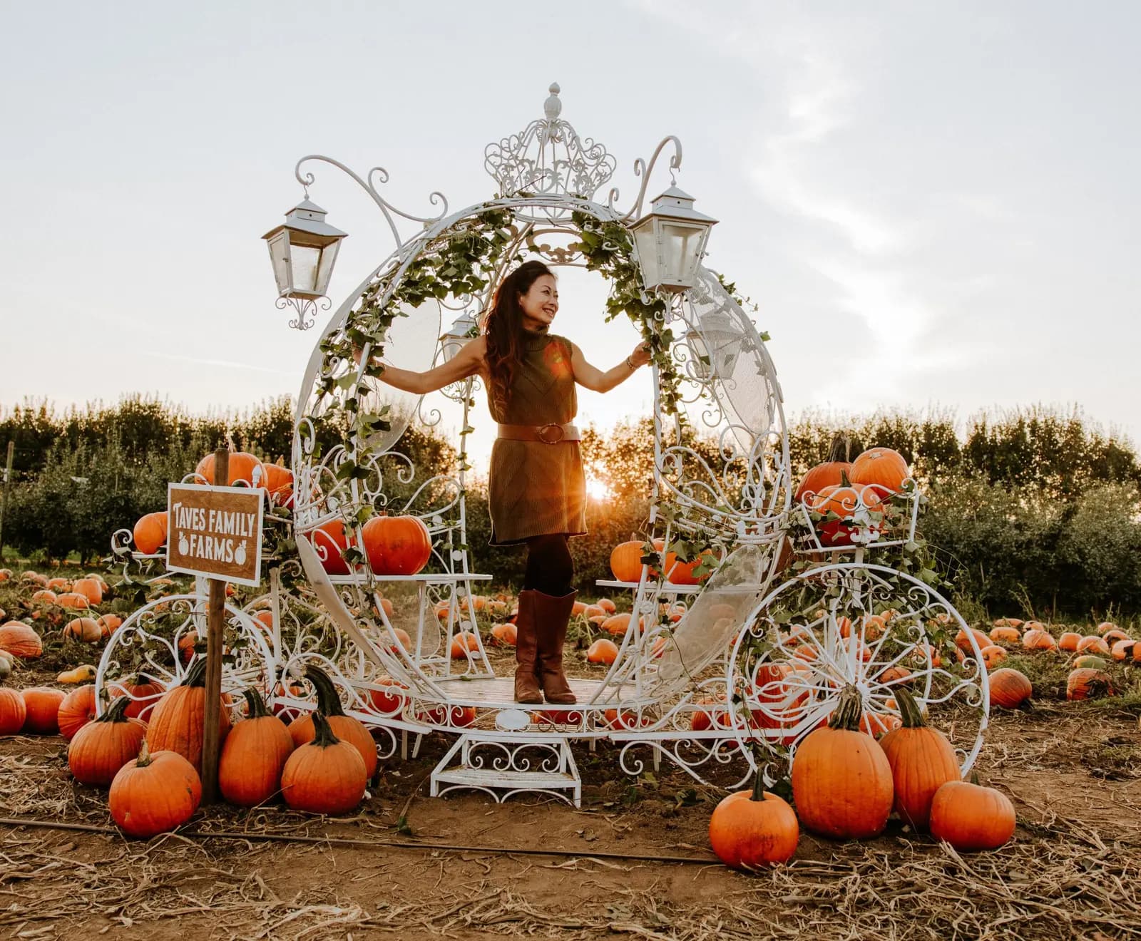 A person stands inside a decorative white carriage frame surrounded by pumpkins at Taves Family Farms.