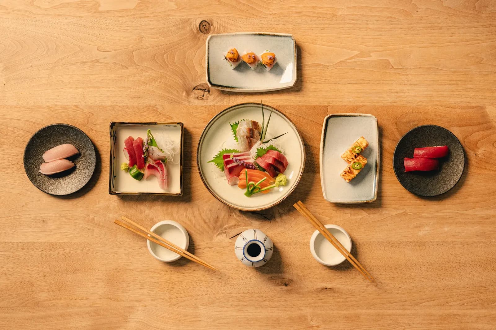 Overhead view of nigiri, sashimi and aburi plates with chopsticks and sake cups on a pale wood table, from Zumi Revolving Sushi Bar